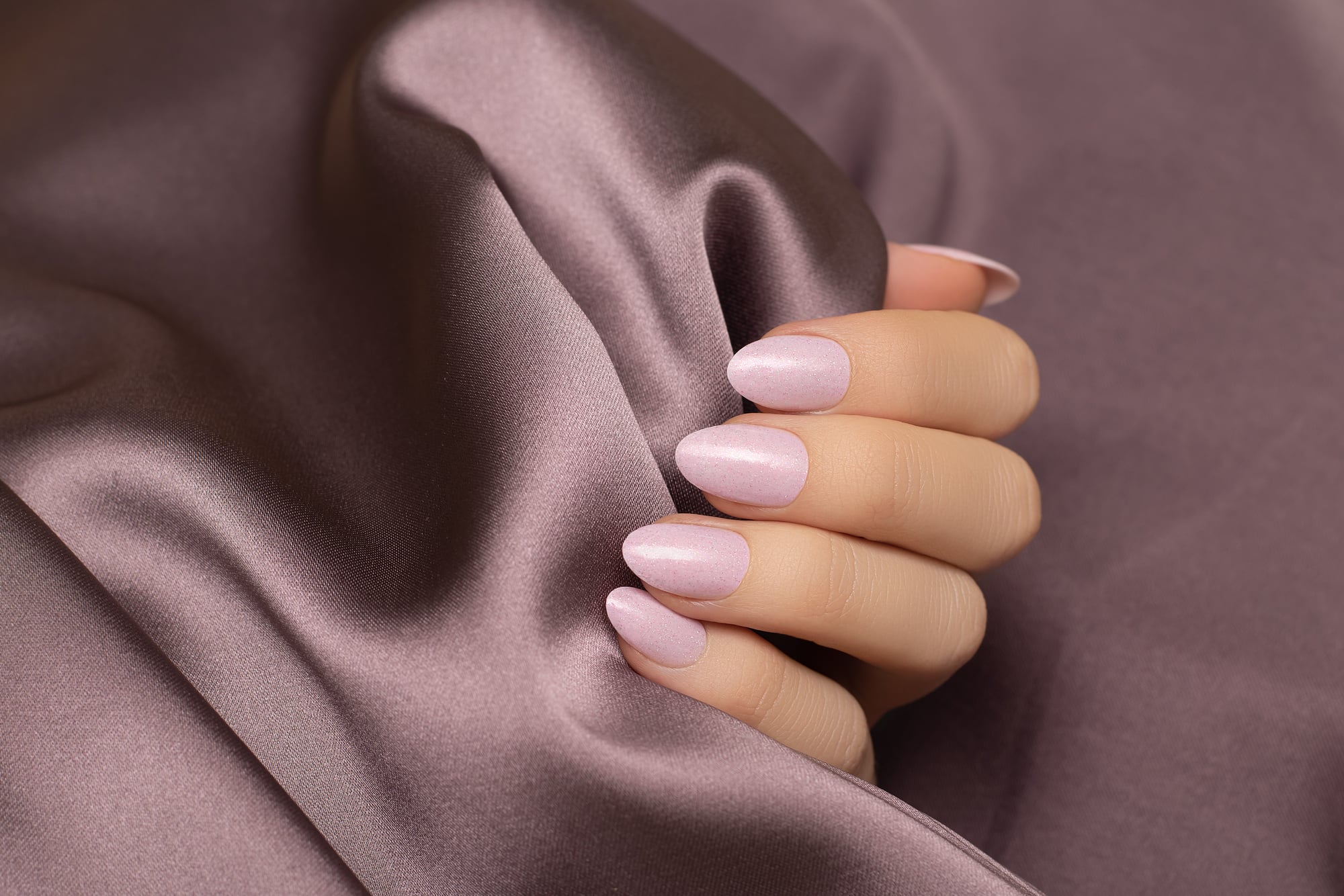 Female hand with pink nail design. Rose glitter nail polish manicure on purple fabric background